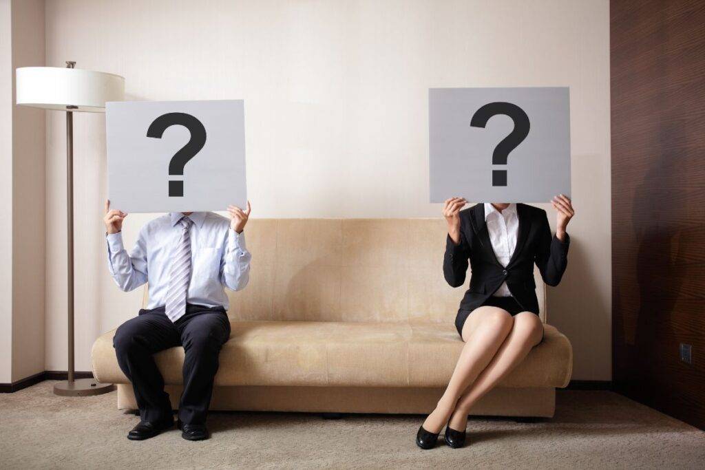 Man and woman sitting on a couch. Each are holding a large question mark over their face.