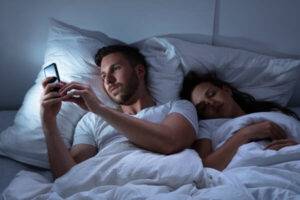 couple in bed - woman is sleeping and man is on his cell phone
