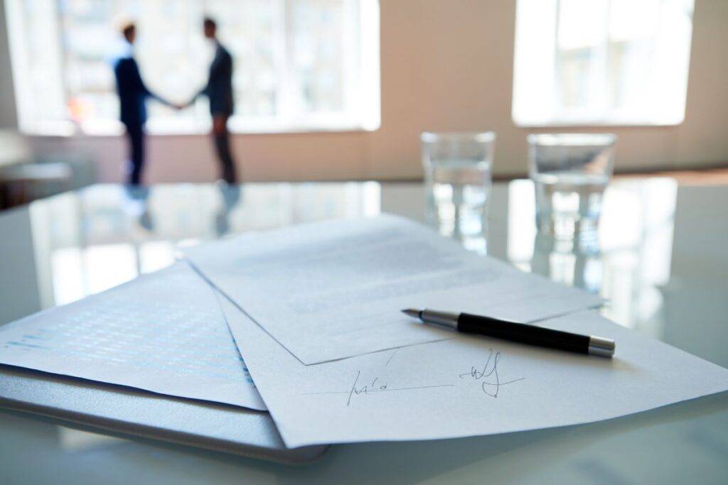 Man in background shaking hands. Papers in foreground are signed.