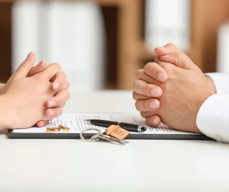 Two pair of hands on the table and facing each other. There  are keys, wedding rings and pen sitting on an annulment or divorce document. 